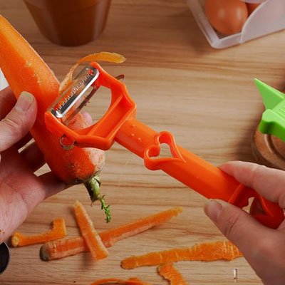 Éplucheur à légumes orange au design original avec poignée en forme de cœur, utilisé pour éplucher une carotte sur une surface en bois