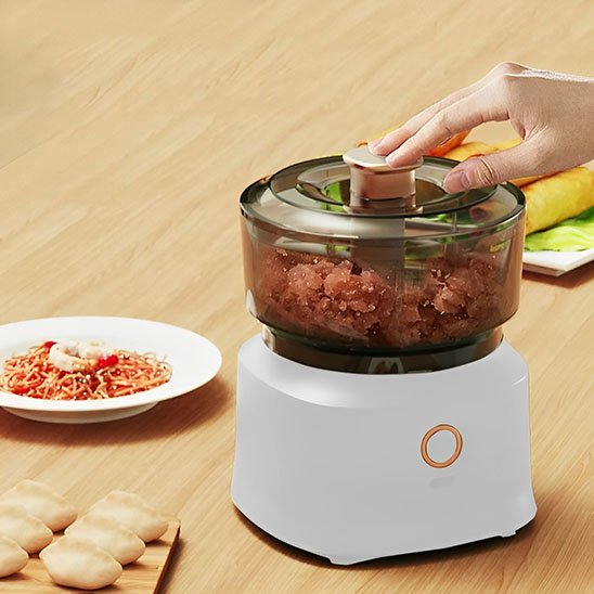 Utilisation d’un hachoir à viande pour préparer de la farce pour raviolis chinois maison sur une table de cuisine avec nouilles et rouleaux en arrière-plan.