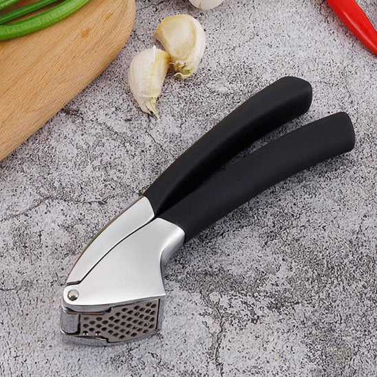  Presse-ail en Inox avec poignées noires sur surface grise, entouré d’ail, légumes verts et piment rouge.