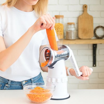 Jeune femme râpant une carotte avec une râpe à légumes manuelle dans une cuisine lumineuse. Les carottes râpées tombent dans un bol en verre, idéales pour une salade fraîche ou une recette maison.
